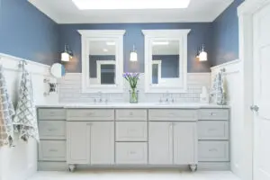 Modern bathroom with double sinks, gray cabinets, white subway tile backsplash, two mirrors, wall sconces, and a vase of purple flowers on the countertop.