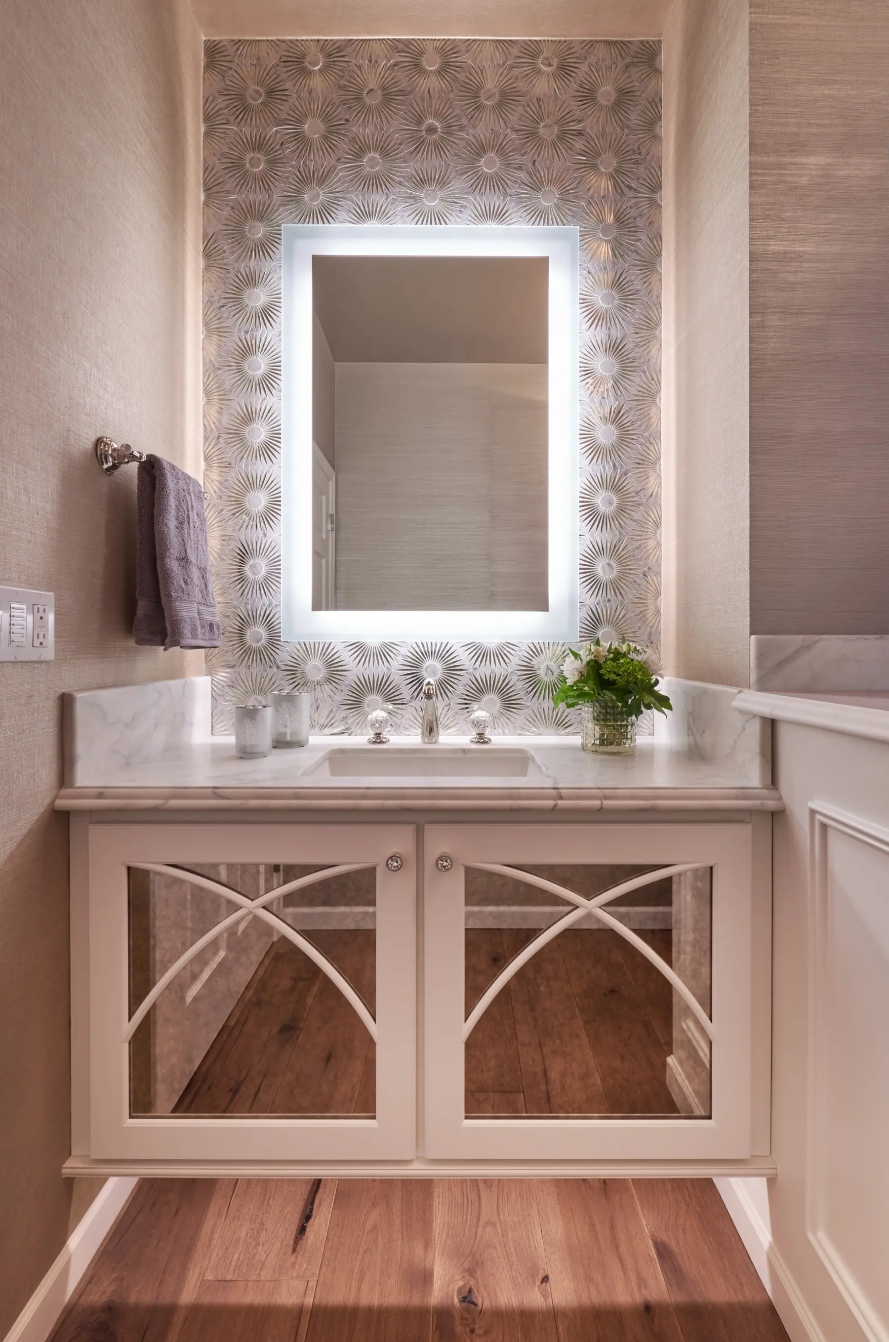 A bathroom vanity with mirrored cabinet doors, a marble countertop, a backlit rectangular mirror, a potted plant, and patterned wallpaper on the wall behind the sink.