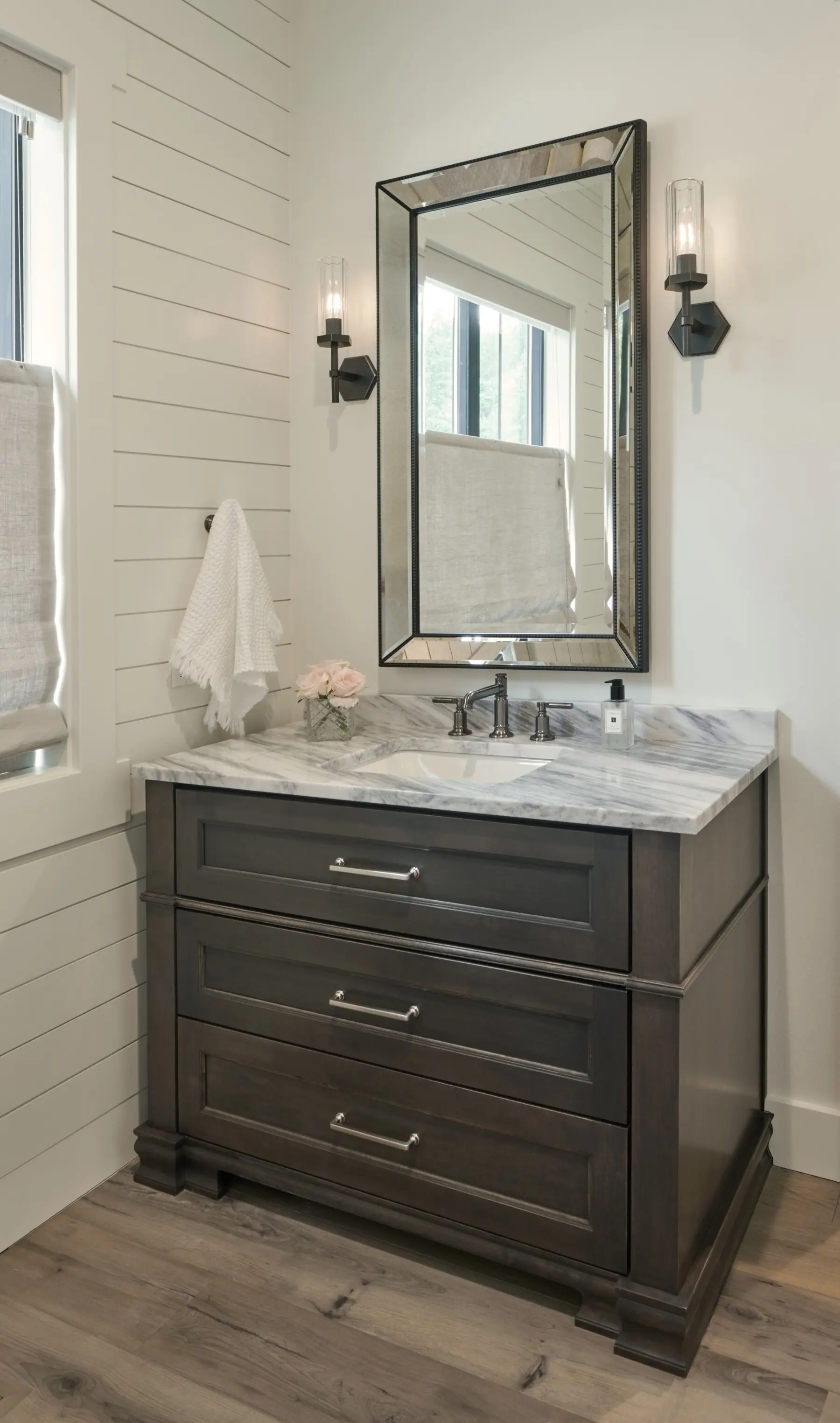 A bathroom vanity with a marble countertop, dark wood drawers, a mirror above, wall sconces on each side, a towel, pink flowers, and a window with a fabric shade.