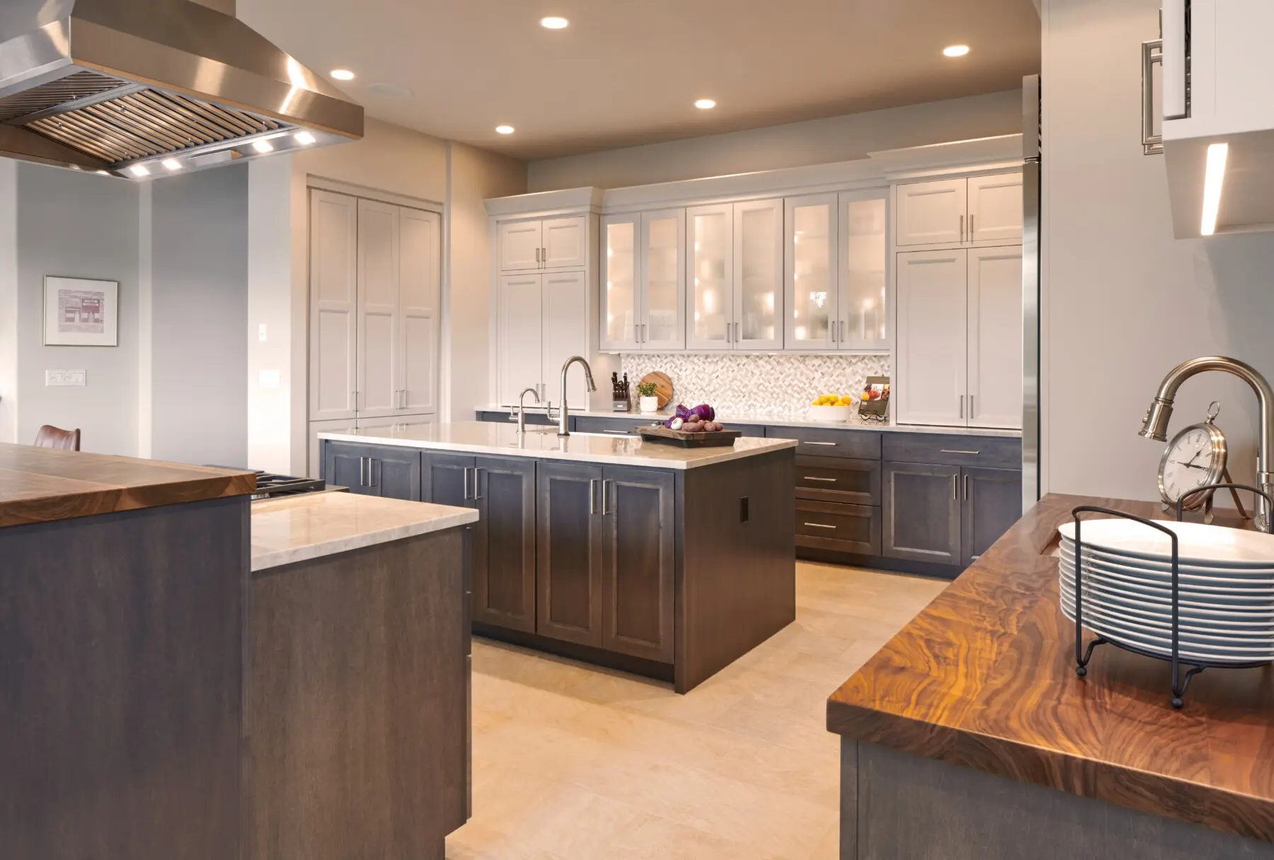 Modern kitchen with two islands, gray cabinets, wood countertops, stainless steel appliances, and under-cabinet lighting. Plates are stacked near a sink on the right.