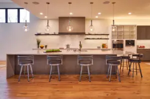 Modern kitchen with a large island, five bar stools, pendant lights, wood flooring, open shelving, built-in appliances, and a white tile backsplash.