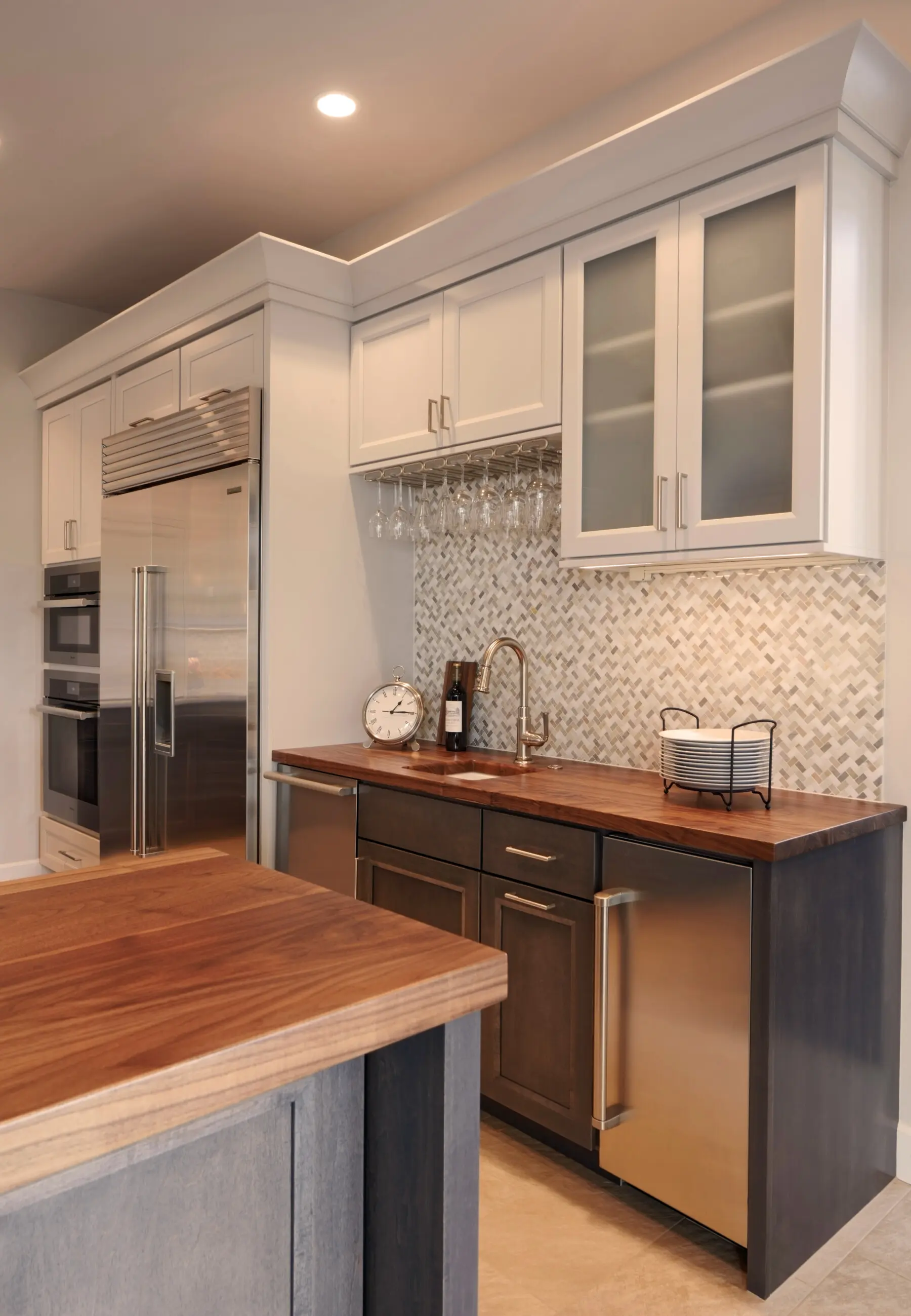 Modern kitchen with stainless steel appliances, wood countertops, glass-front cabinets, backsplash tile, wine glasses hanging, and under-cabinet sink area.