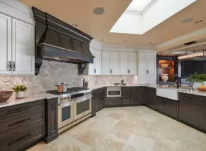 Modern kitchen with dark lower cabinets, white upper cabinets, stainless steel appliances, a farmhouse sink, and beige tiled floor under a skylight.