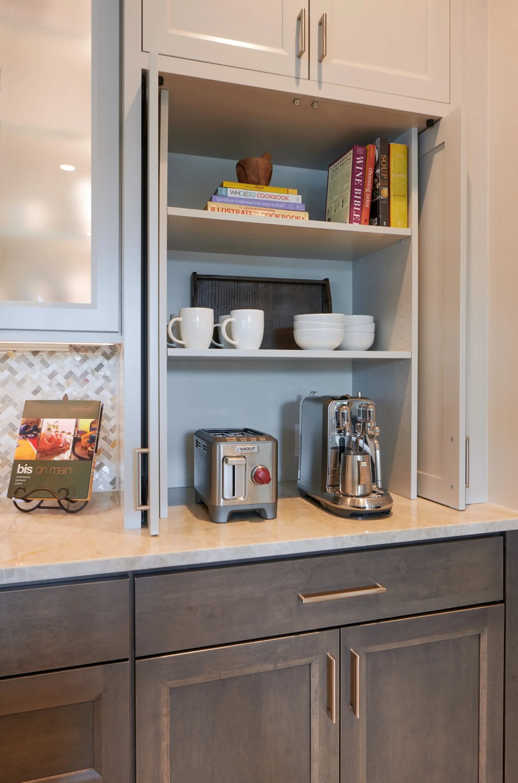 A kitchen cabinet with open doors displays cups, bowls, books, a toaster, and a coffee maker on shelves above a beige countertop with gray cabinets below.