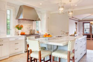 Modern kitchen with white cabinets, a marble island with seating, stainless steel range hood, pendant lights, and a bowl of oranges on the counter.