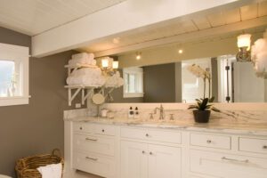 Modern bathroom with white cabinets, marble countertop, large mirror, rolled towels on shelves, a potted orchid, and a wicker basket on the floor.