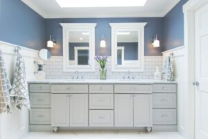 Modern bathroom with double sinks, gray cabinets, white subway tile backsplash, two mirrors, wall sconces, and a vase of purple flowers on the countertop.