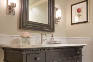 A bathroom vanity with a marble countertop, a framed mirror, wall sconces, a flower vase, soap dispenser, and a framed flower picture on the wall.