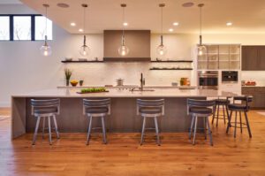 Modern kitchen with a large island, five bar stools, pendant lights, wood flooring, open shelving, built-in appliances, and a white tile backsplash.