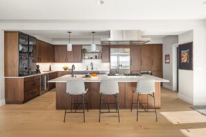 Modern kitchen with wooden cabinets, stainless steel appliances, a large island with three gray barstools, pendant lights, and light wood flooring.