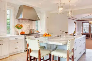 Modern kitchen with white cabinets, a marble island with seating, stainless steel range hood, pendant lights, and a bowl of oranges on the counter.