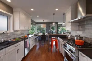 Modern kitchen with white cabinets, gray backsplash, stainless steel appliances, a breakfast nook with large windows, and wooden flooring. Orange barstool and cookware add color accents.