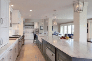 Spacious modern kitchen with white cabinets, marble countertops, pendant lighting, and a view into a living area with large windows. Fruit bowl on the central island.