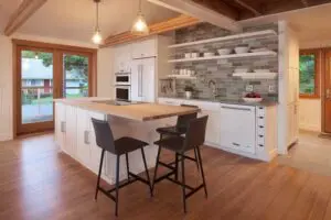 Modern kitchen with wood floors, white cabinets, a center island with two black chairs, open shelves, and gray tile backsplash. Large windows and pendant lights provide natural and artificial light.