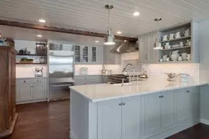 Modern kitchen with gray cabinets, a large white island countertop, stainless steel appliances, open shelving with dishes, and pendant lights over a dark wood floor.