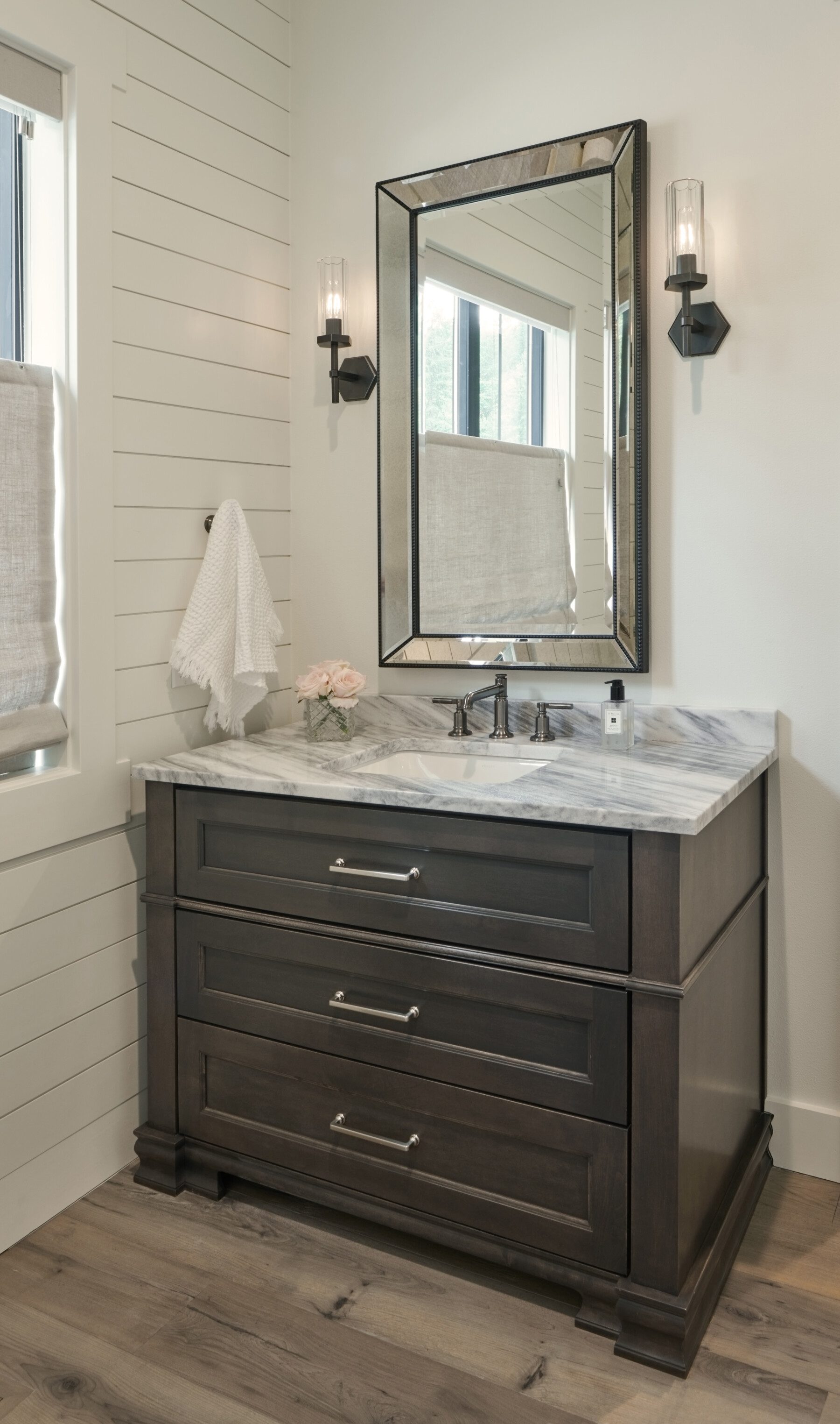 A bathroom vanity with a marble countertop, dark wood drawers, a mirror above, wall sconces on each side, a towel, pink flowers, and a window with a fabric shade.