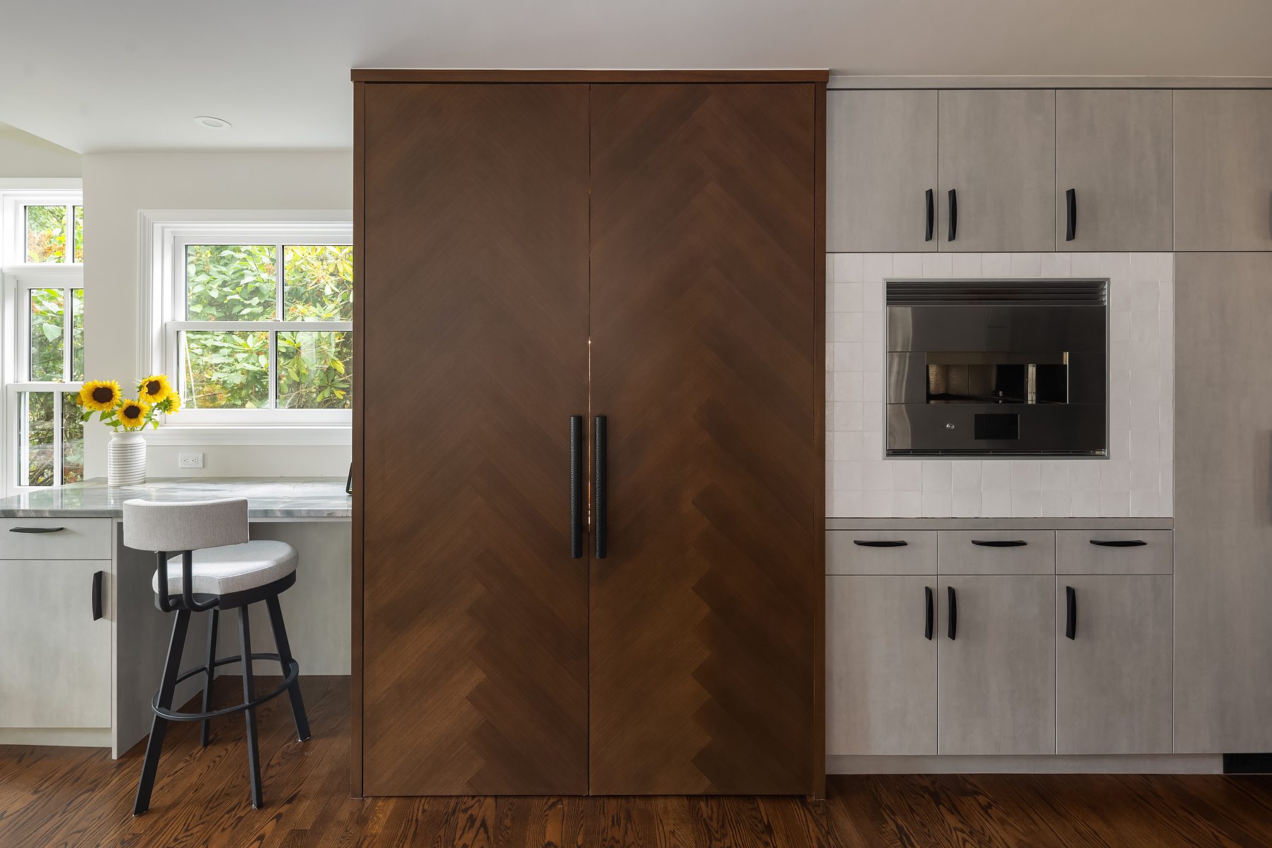 Modern kitchen with a large wooden cabinet in the center, gray cabinetry with black handles, a built-in microwave, and a countertop with sunflowers in a vase by the window.