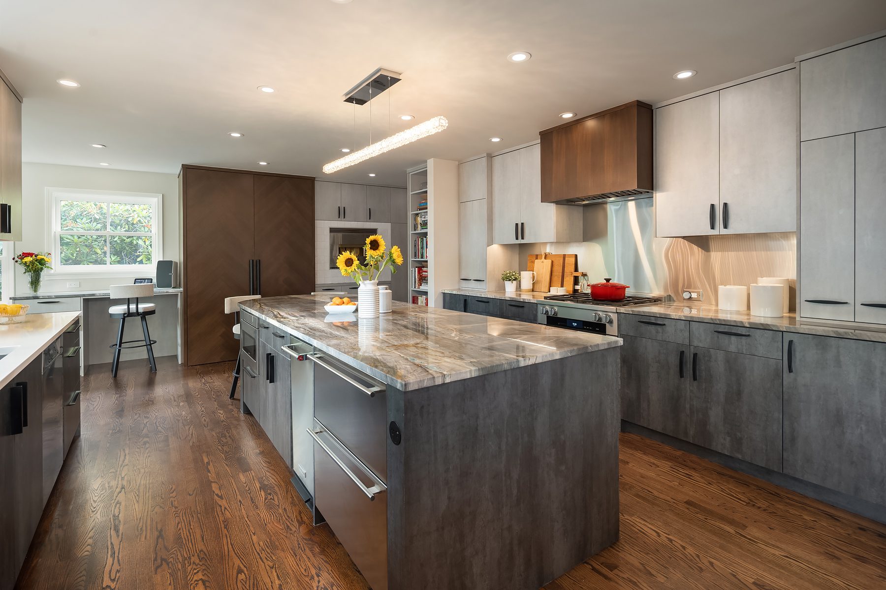Modern kitchen with gray cabinets, marble countertops, a large center island, wood flooring, sunflowers in a vase, and under-cabinet lighting.