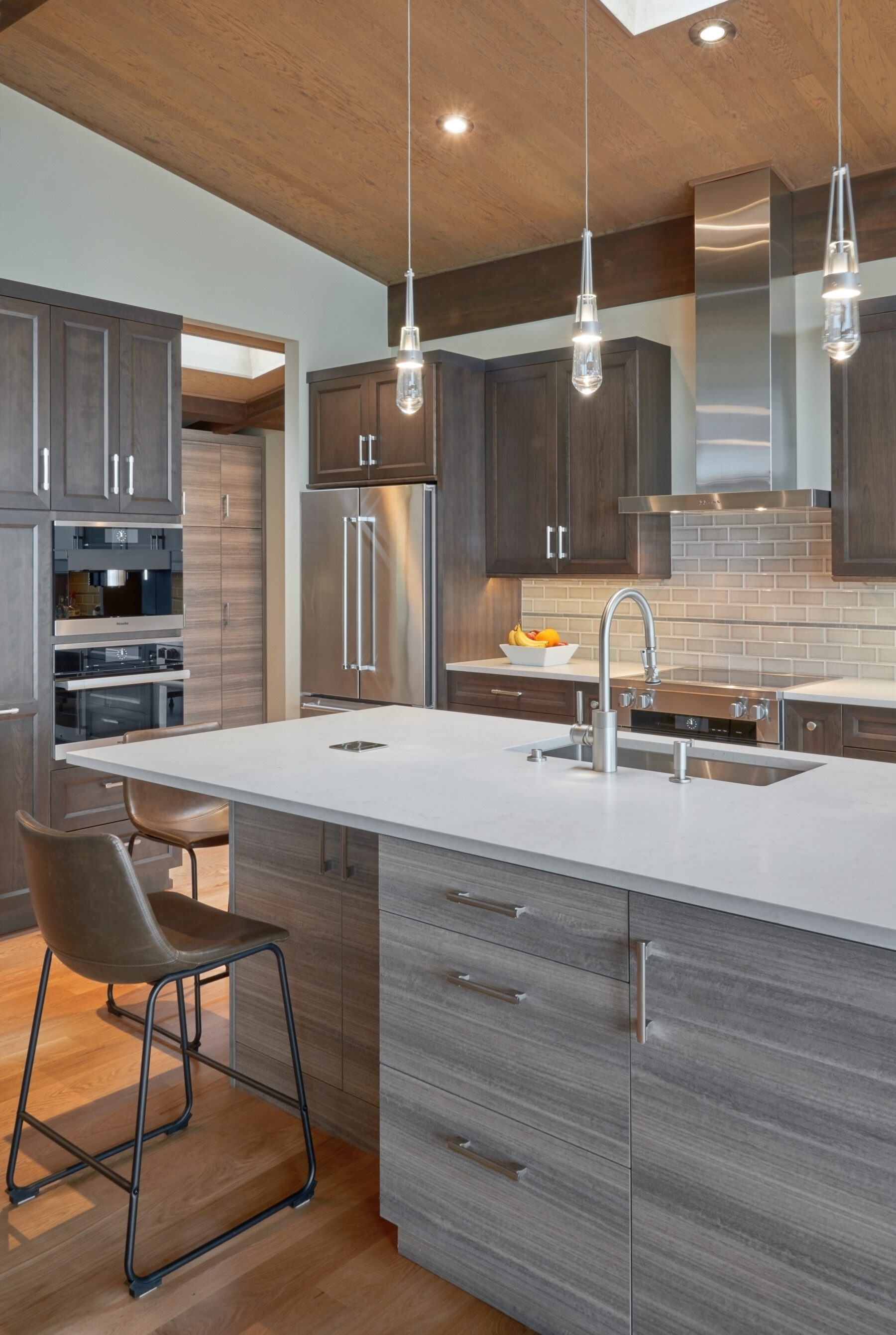 Modern kitchen with gray wood cabinets, stainless steel appliances, pendant lights, a white island countertop, and a brown bar stool on a wooden floor.