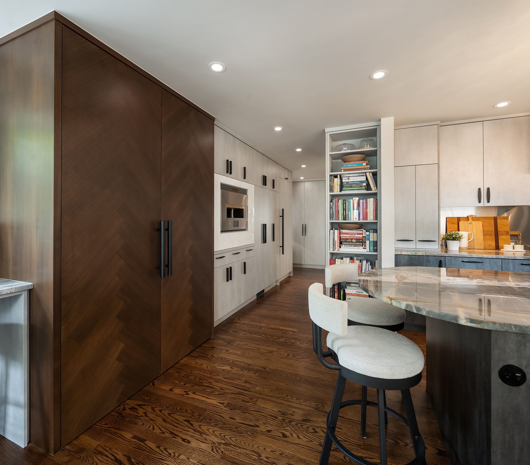 Modern kitchen with wood flooring, light and dark cabinetry, a marble countertop island with two cushioned bar stools, built-in appliances, and a tall bookshelf filled with cookbooks.