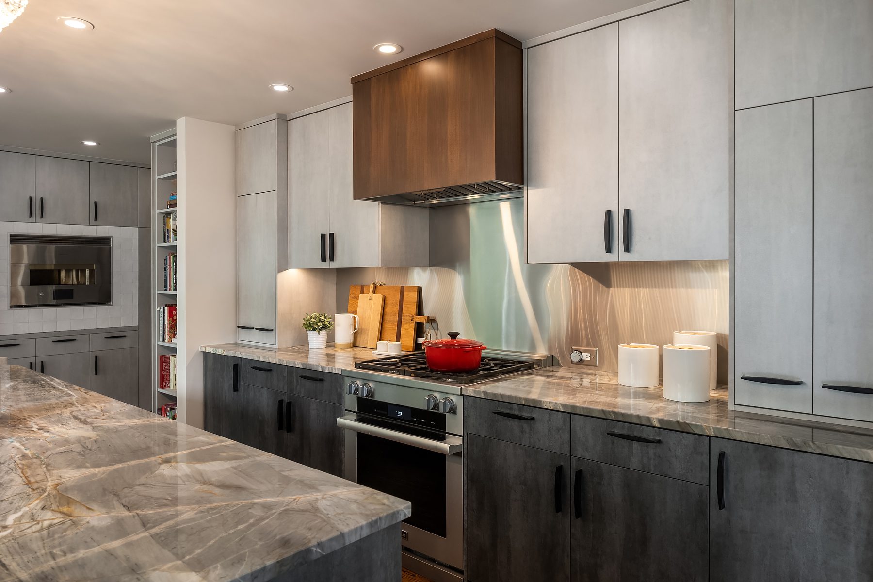 Modern kitchen with gray cabinets, marble countertops, stainless steel appliances, a red pot on the stove, under-cabinet lighting, and various kitchen accessories on the counters.