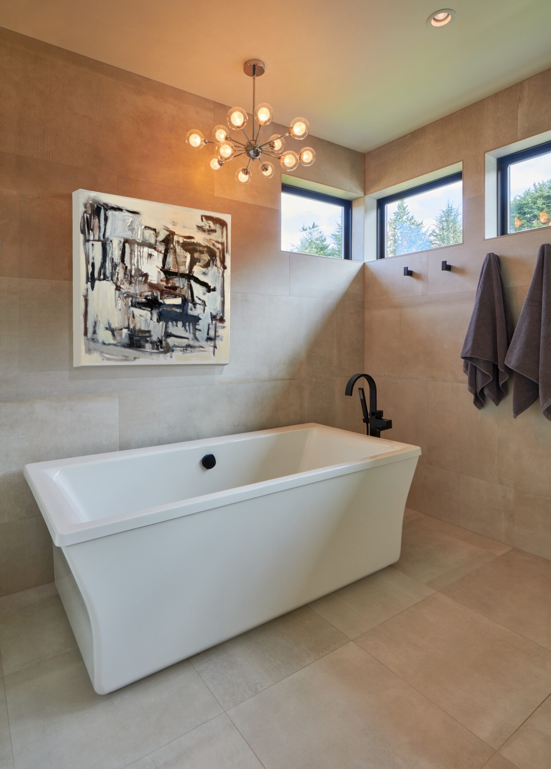 Modern bathroom with a white freestanding bathtub, wall-mounted black faucet, two gray towels, abstract wall art, and a contemporary light fixture.