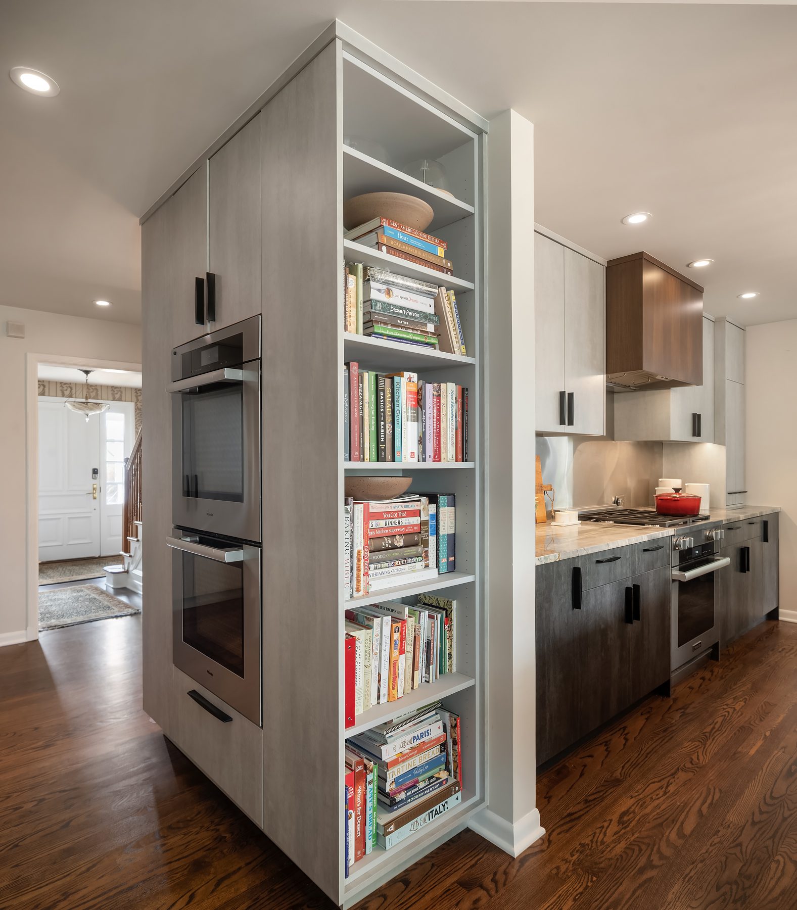 Modern kitchen with built-in ovens and a tall bookshelf filled with cookbooks, featuring wood floors, gray cabinets, and stainless steel appliances.