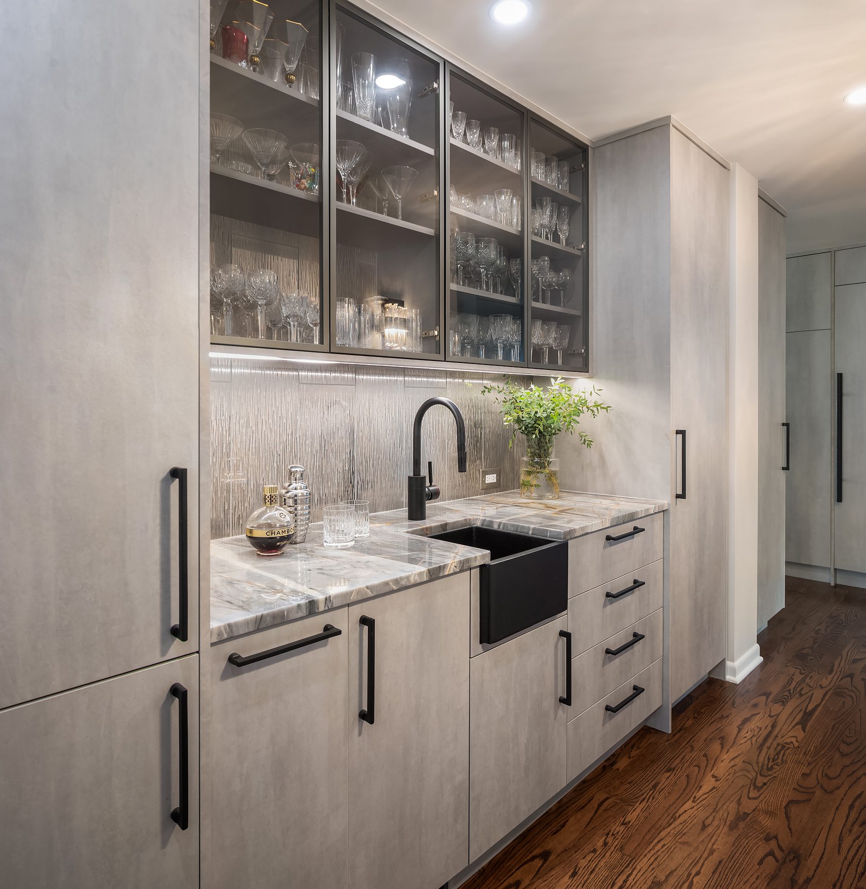 Modern kitchen with light gray cabinets, glass-front upper shelves displaying glassware, a black faucet, marble countertop, and wooden flooring. A small plant sits near the sink.