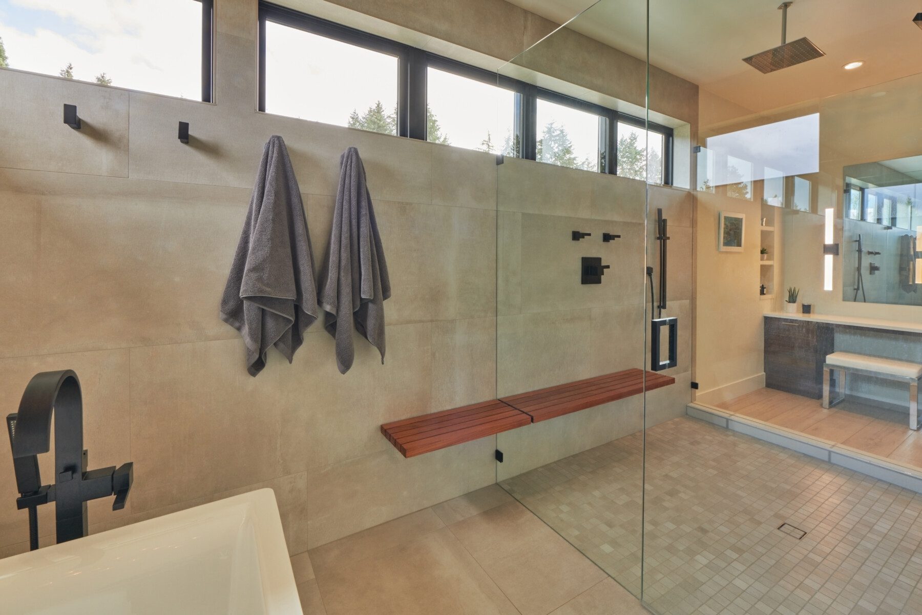 Modern bathroom with a glass-enclosed shower, wood bench, wall-mounted fixtures, two gray towels, and a bathtub; large windows provide natural light.