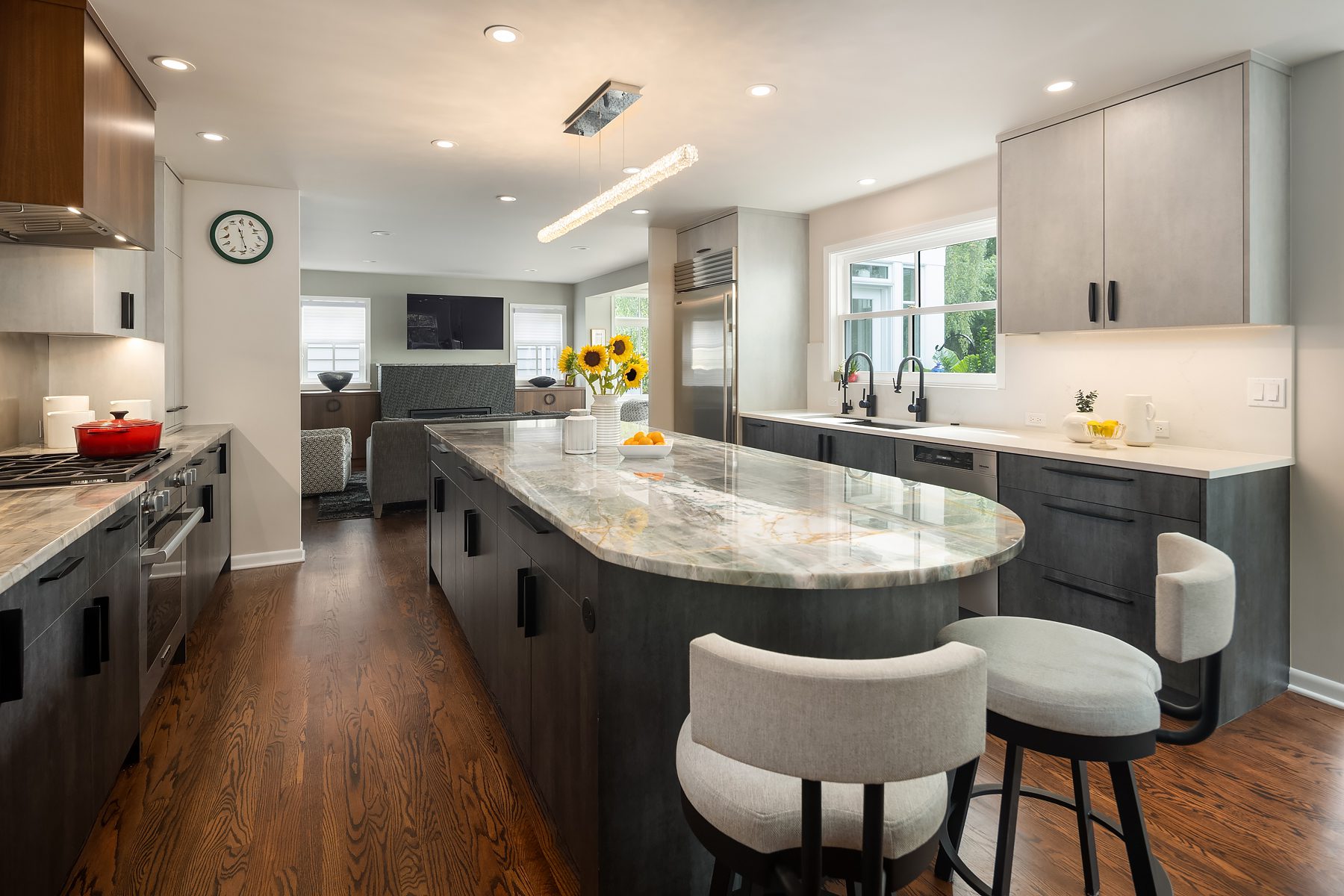 Modern kitchen with a large marble island, gray cabinets, stainless steel appliances, bar stools, sunflowers in a vase, and hardwood floors, with a view into the living area.