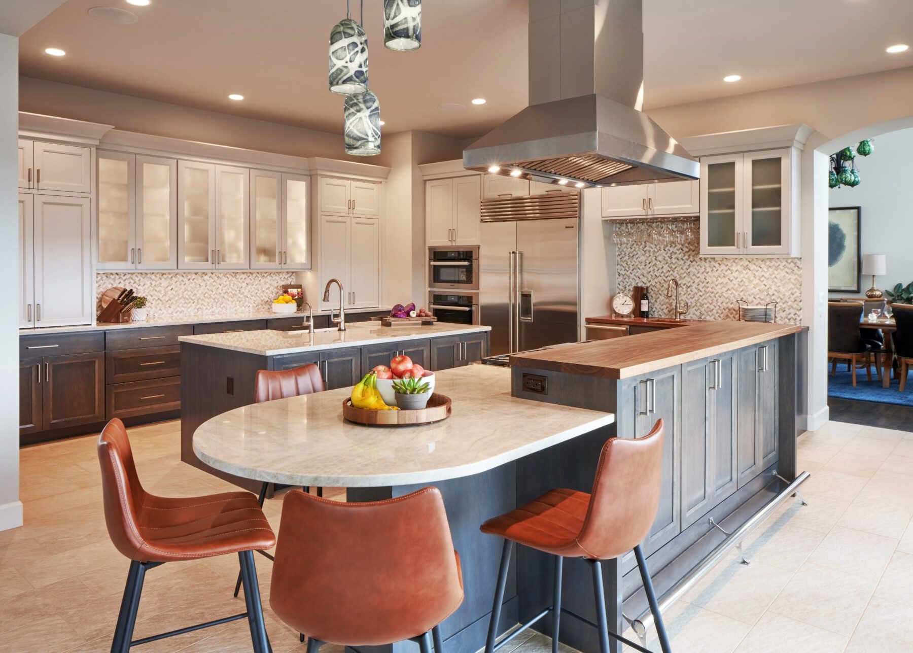 Contemporary kitchen with stainless steel appliances, marble countertops, brown cabinets, and four brown leather bar stools around a curved island. Fruit bowl on island, dining area in background.