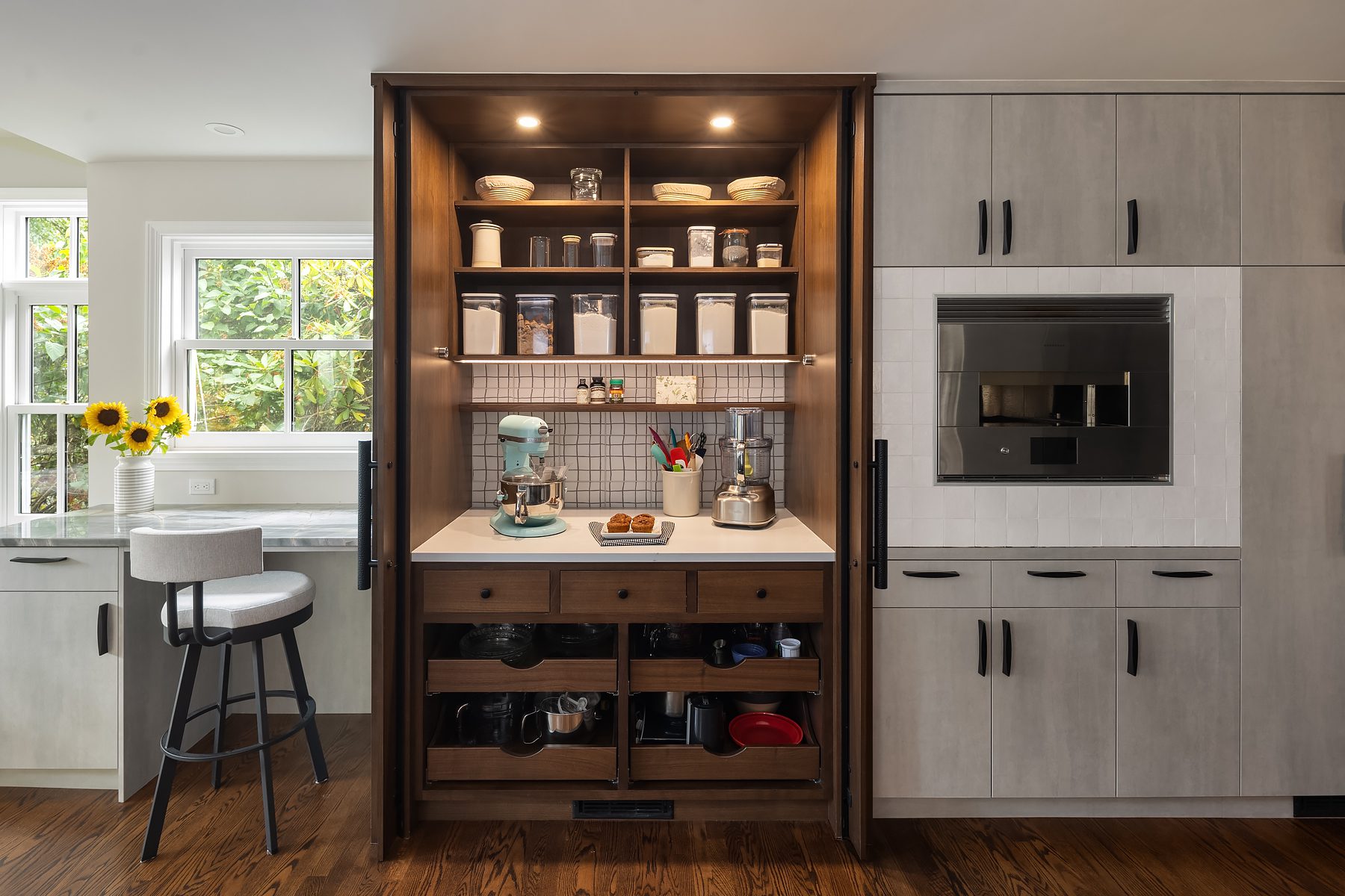 A modern kitchen features an open pantry with shelves holding jars, dishes, and appliances, flanked by closed cabinets and a built-in oven. A stool sits near a counter with sunflowers.