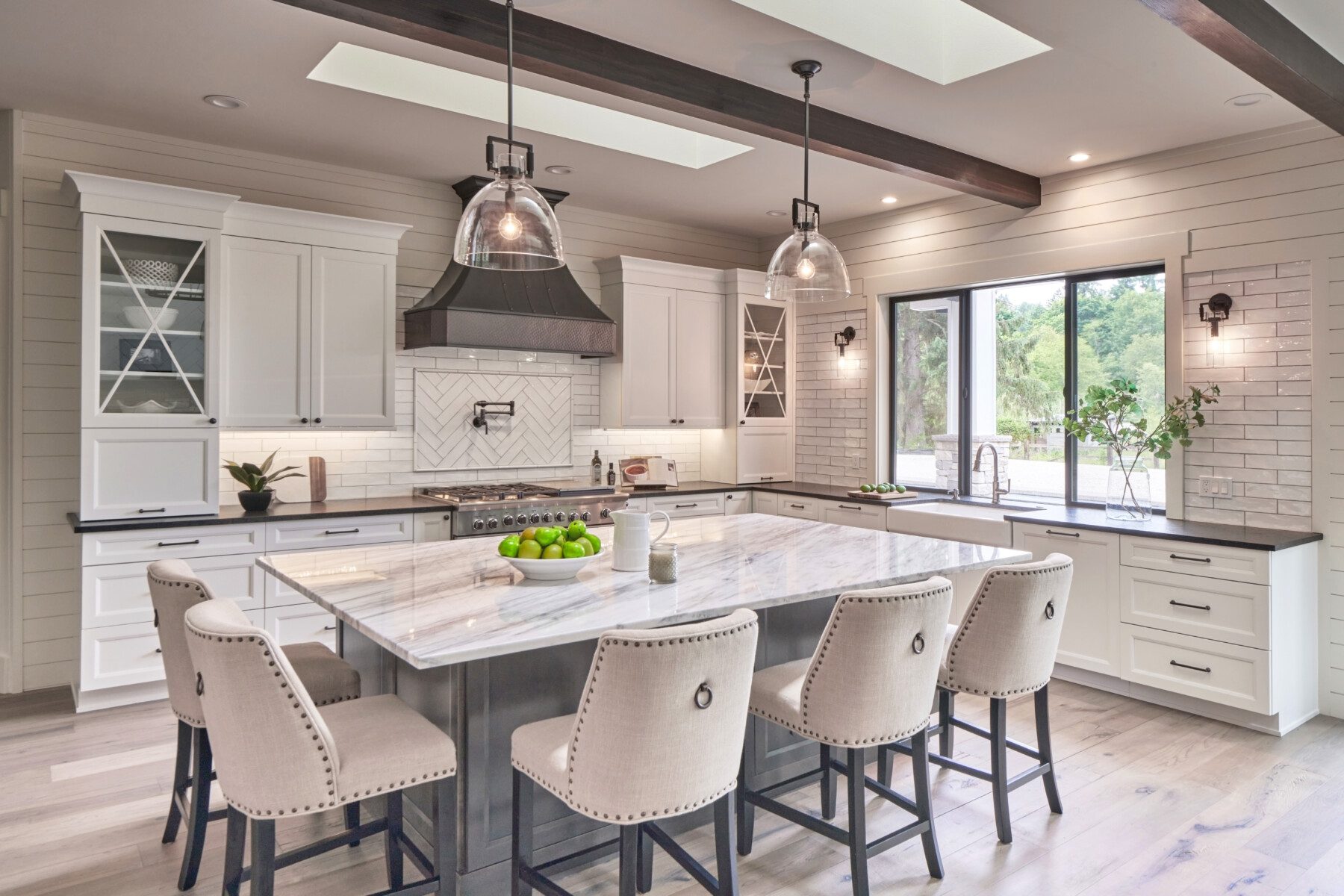 Modern kitchen with white cabinets, marble island, upholstered chairs, pendant lights, stainless steel appliances, and large windows overlooking greenery.