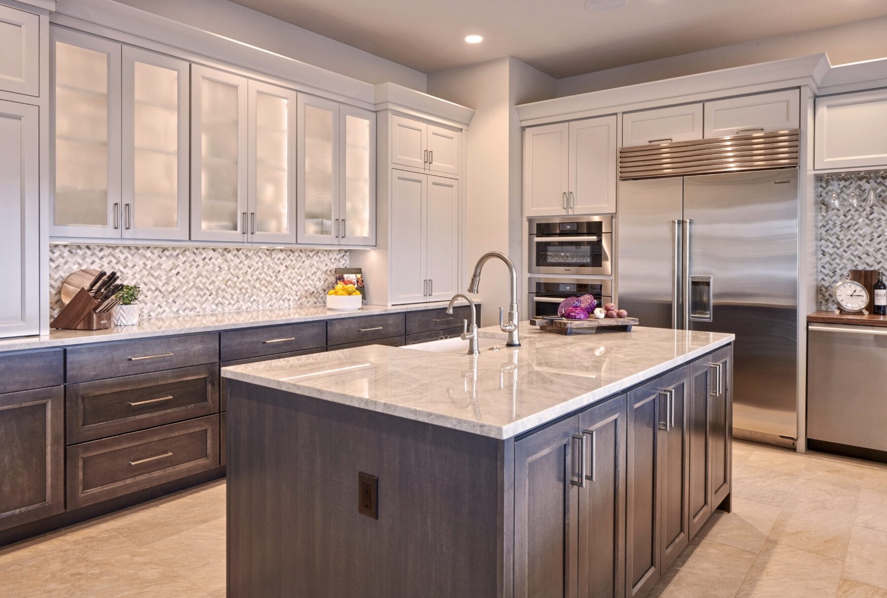 Modern kitchen with a large island, marble countertops, stainless steel appliances, and glass-front upper cabinets. Cutting boards and fruit are arranged on the counter.