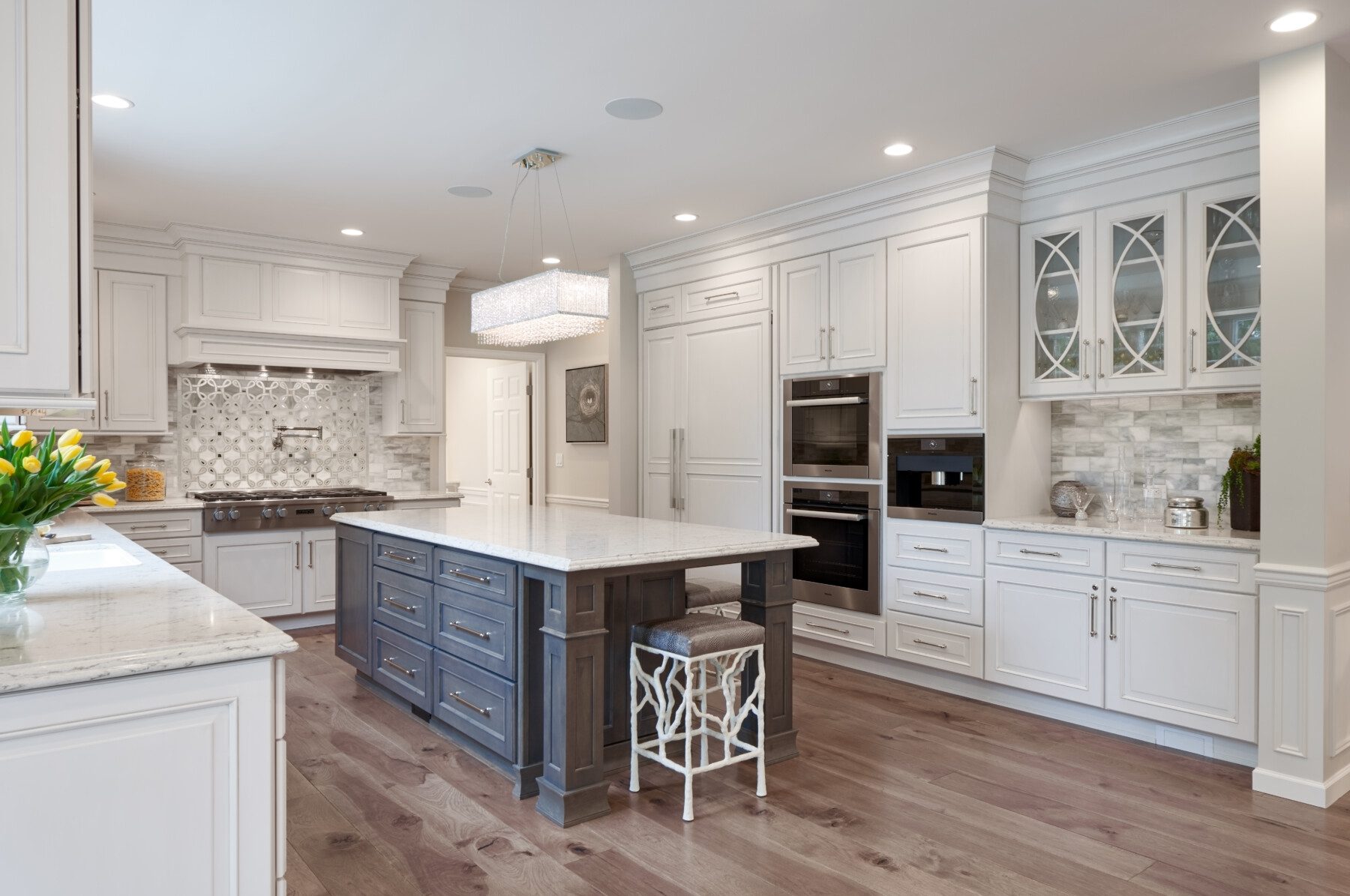 Spacious modern kitchen with white cabinets, stainless steel appliances, a large island with a dark base, marble countertops, and wood flooring.