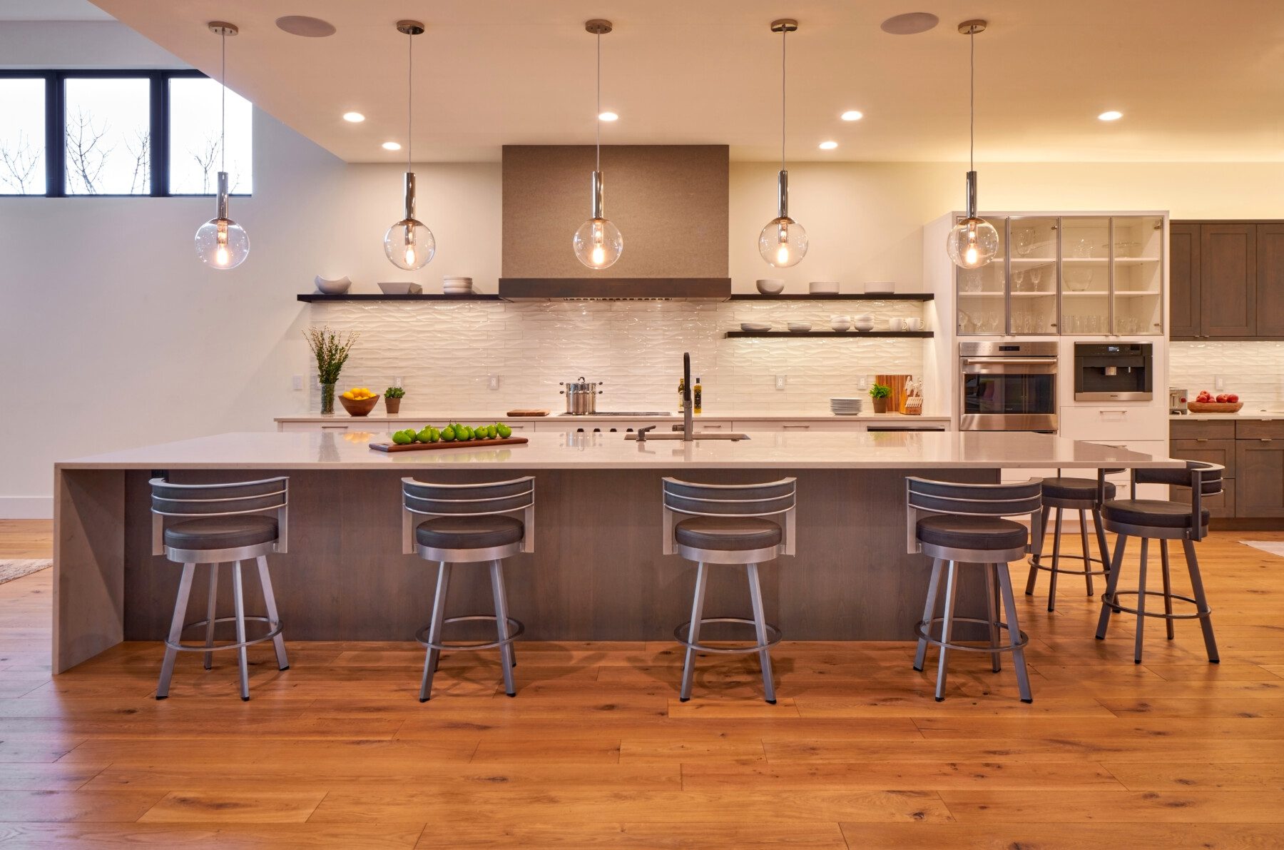 Modern kitchen with a large island, five bar stools, pendant lights, wood flooring, open shelving, built-in appliances, and a white tile backsplash.