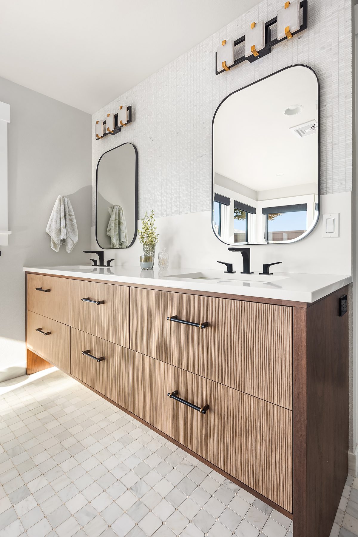 Modern bathroom with a wood vanity, double sinks, two rectangular mirrors, wall-mounted lights, white tile backsplash, and floor tiles. Towels hang on the left wall.