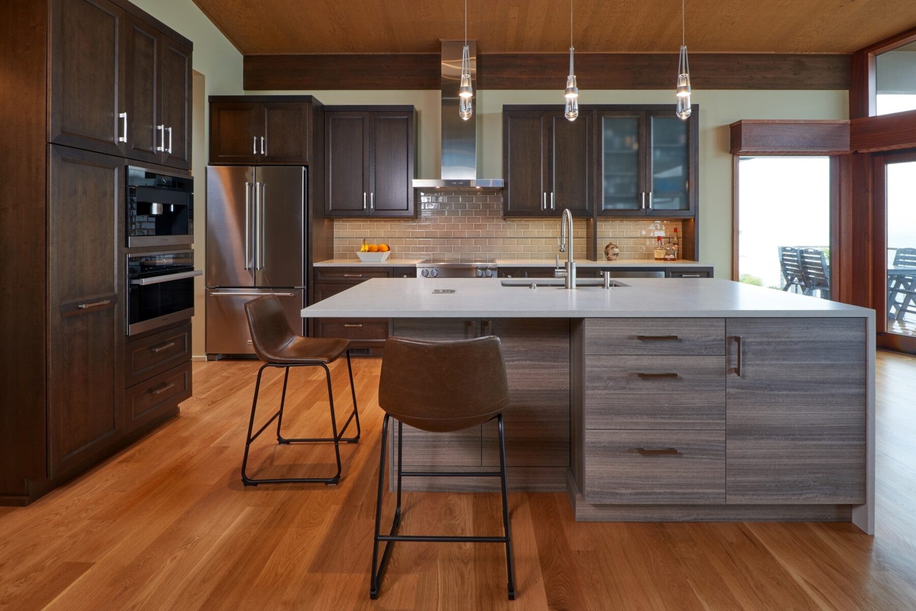 Modern kitchen with dark wood cabinets, stainless steel appliances, a center island with a sink, three pendant lights, and two brown bar stools on a wood floor.
