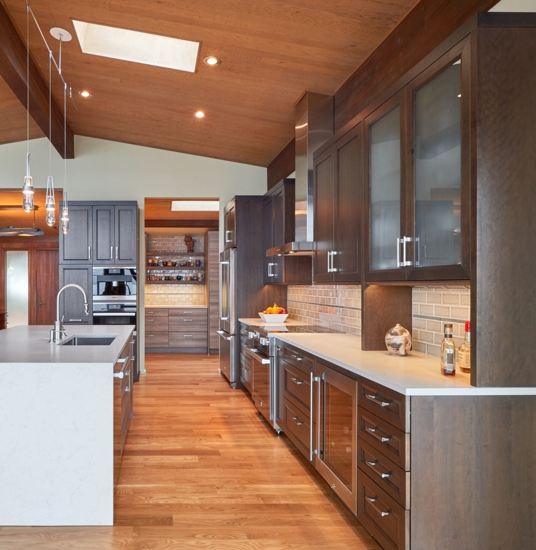 Modern kitchen with wooden floors, dark wood cabinets, stainless steel appliances, a central island with a sink, and pendant lighting under a wood-paneled ceiling.
