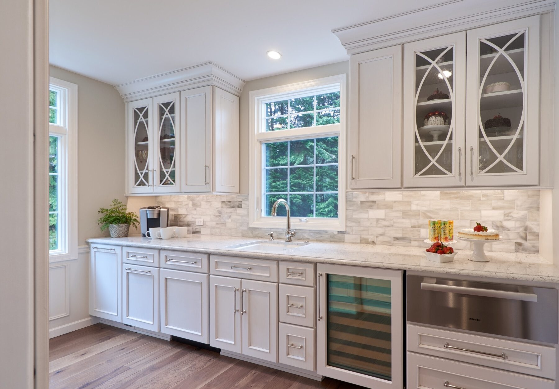Bright kitchen with white cabinets, glass-front doors, marble backsplash, under-counter wine cooler, built-in dishwasher, potted plant, and desserts displayed on the counter.