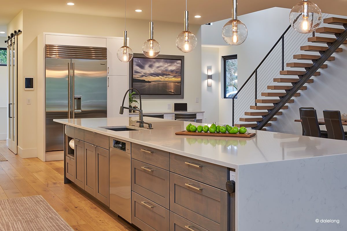 Modern kitchen with a large island, pendant lights, stainless steel appliances, and a tray of green apples; open stairway and dining area in the background.