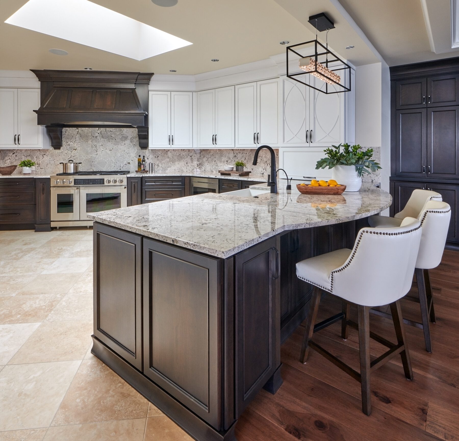 Spacious kitchen with a large marble island, dark and white cabinetry, built-in stove, light tile flooring, and two white bar stools.