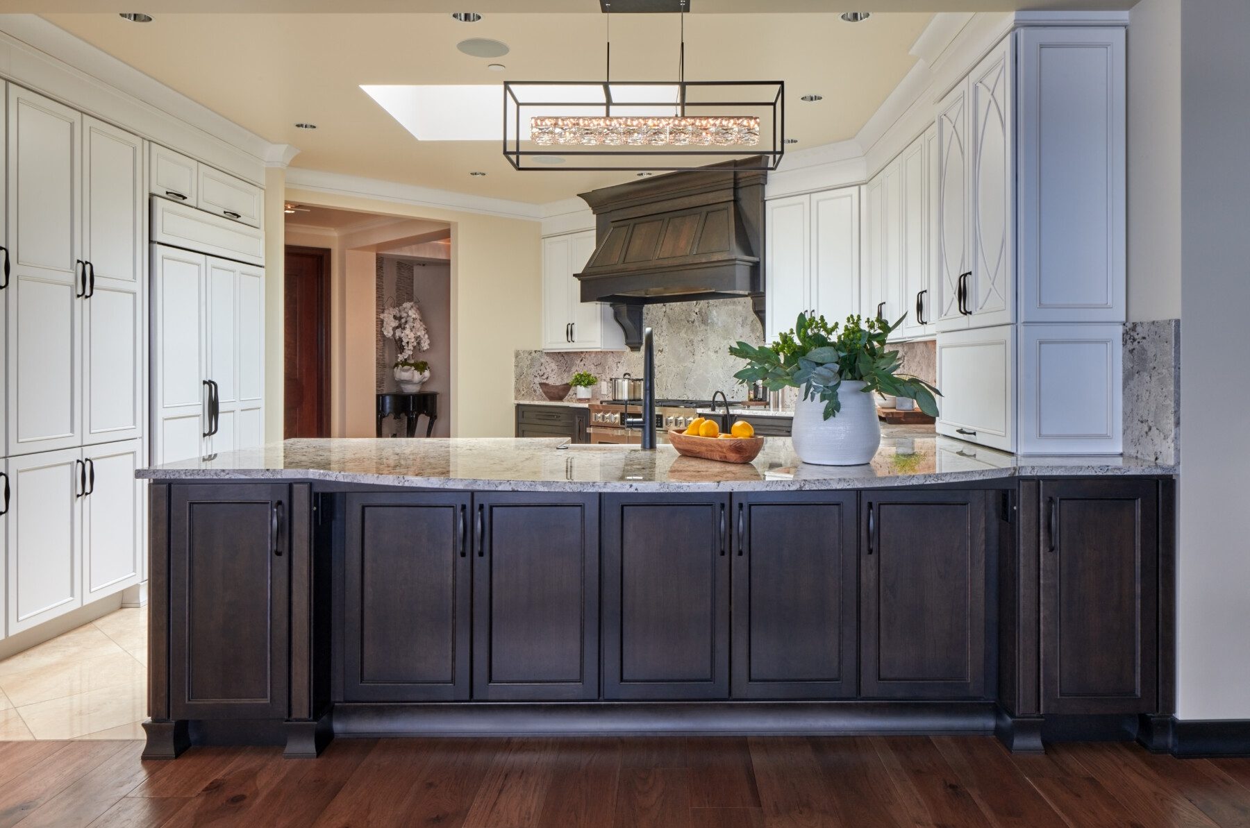 Modern kitchen with dark wood island, white cabinets, marble countertops, stainless steel appliances, and a vase with greenery on the counter. Oranges sit on a wooden cutting board.