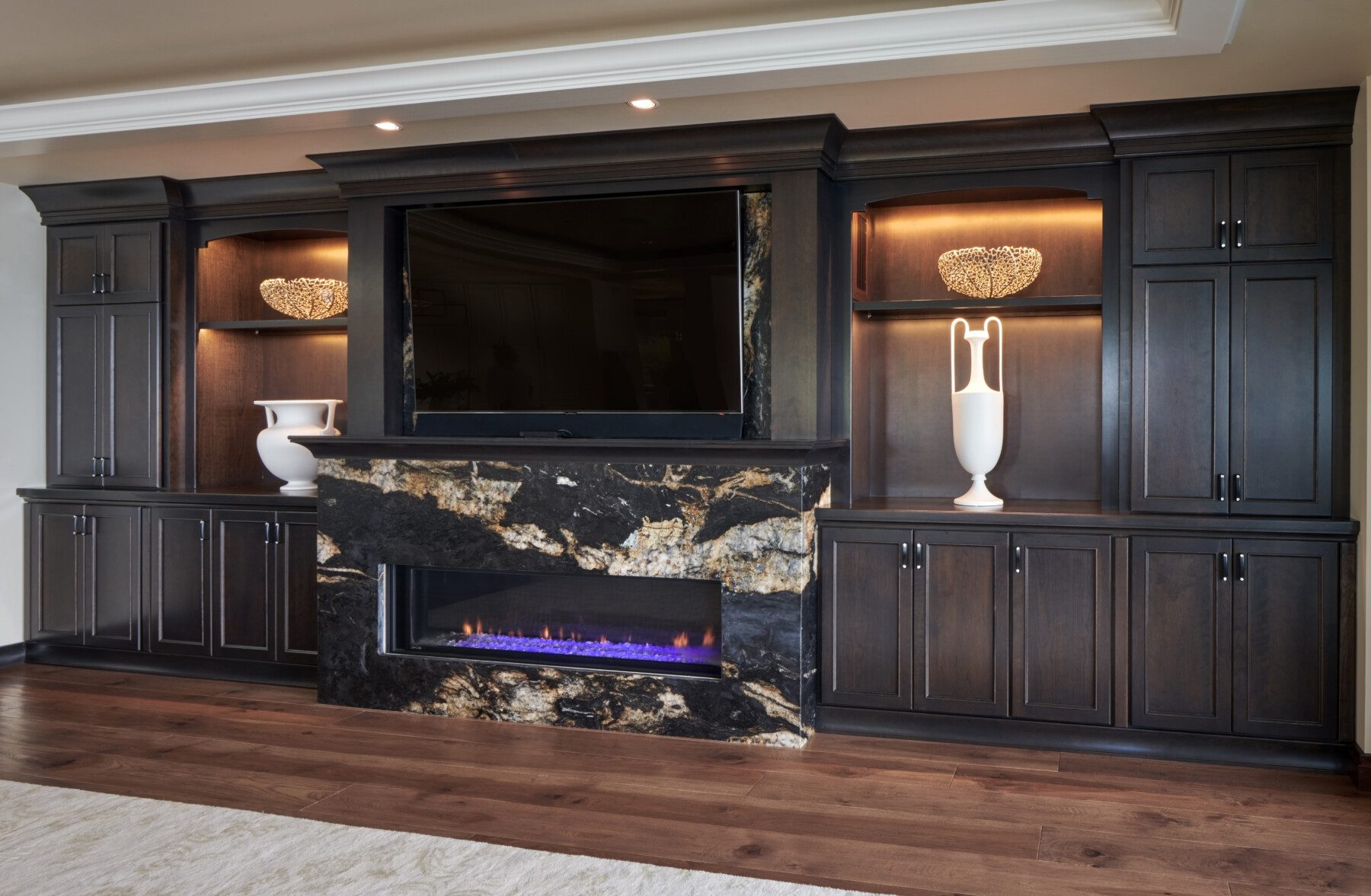 Built-in dark wood entertainment center features a large TV above a marble fireplace with blue flames, shelves displaying white vases, and cabinets below.