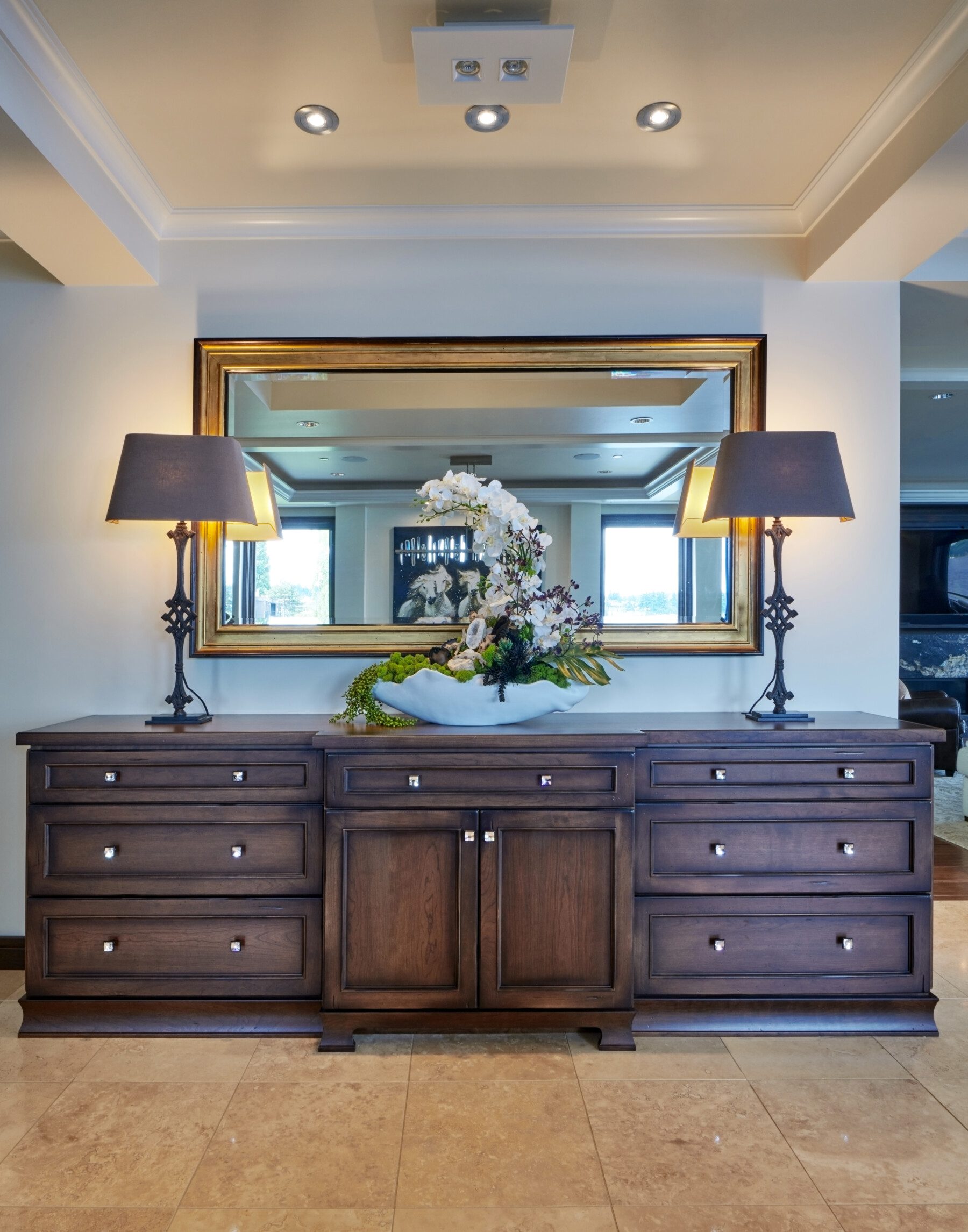 A dark wood sideboard with drawers is topped by two lamps, a decorative bowl with flowers, and stands in front of a large gold-framed mirror in a well-lit room.