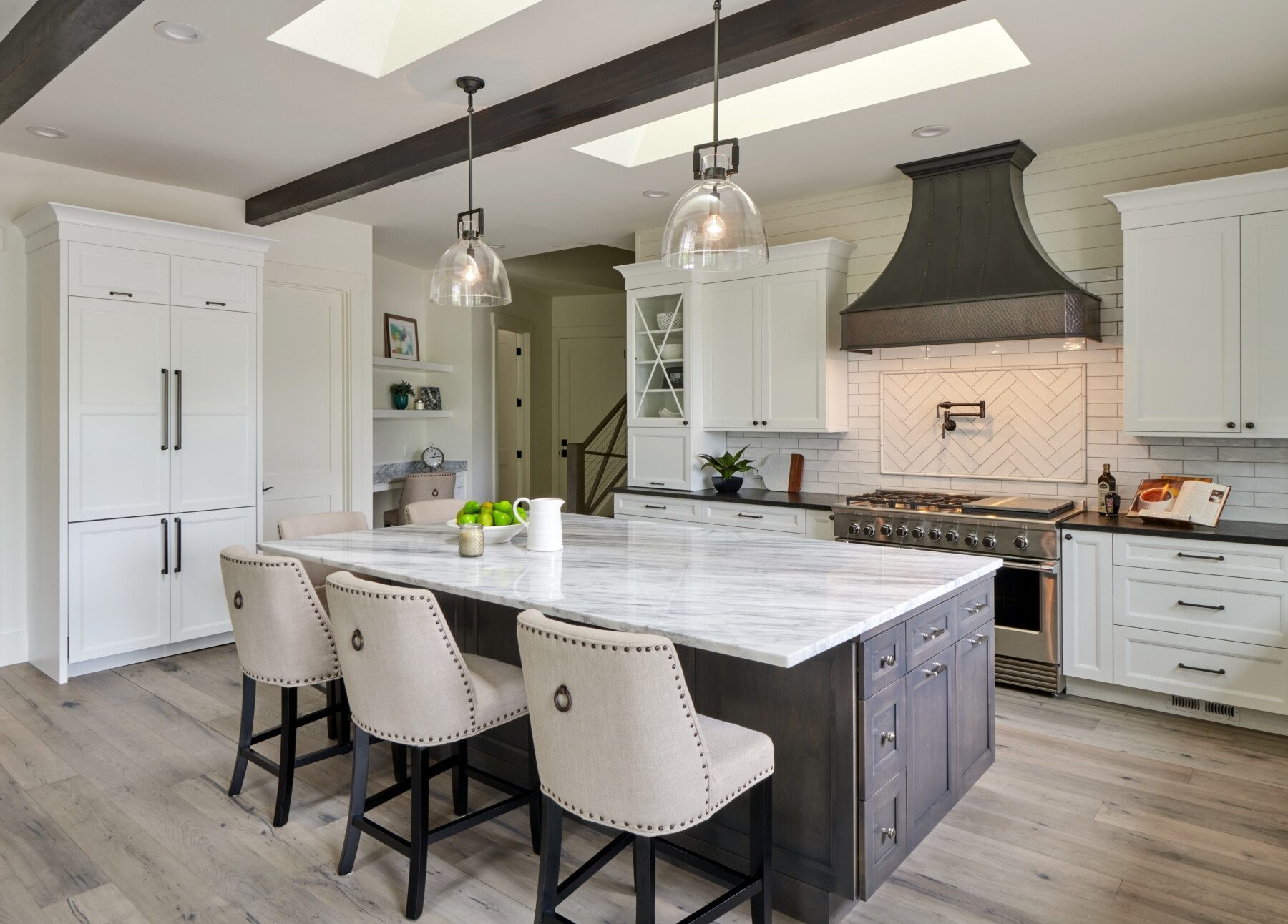 Modern kitchen with a large marble island, four upholstered chairs, white cabinets, stainless steel appliances, wood flooring, and pendant lights under skylights.
