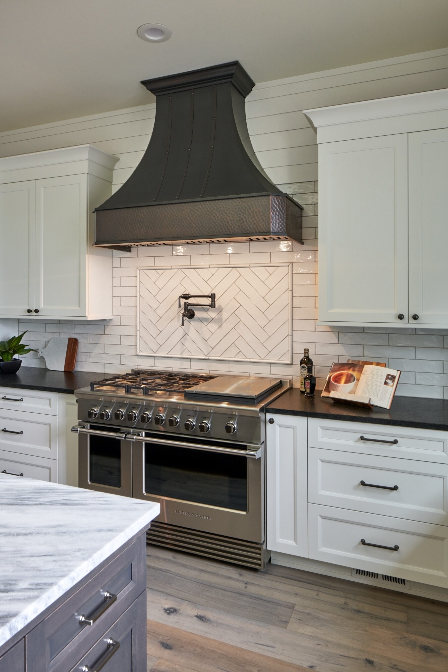 Modern kitchen with a stainless steel gas range, black range hood, white cabinets, herringbone tile backsplash, and an open cookbook on the counter.