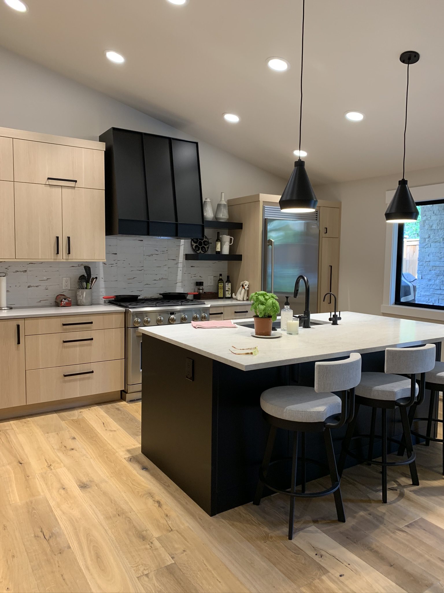 Modern kitchen with light wood cabinets, black island, stainless steel appliances, two bar stools, pendant lights, and a potted plant on the counter.