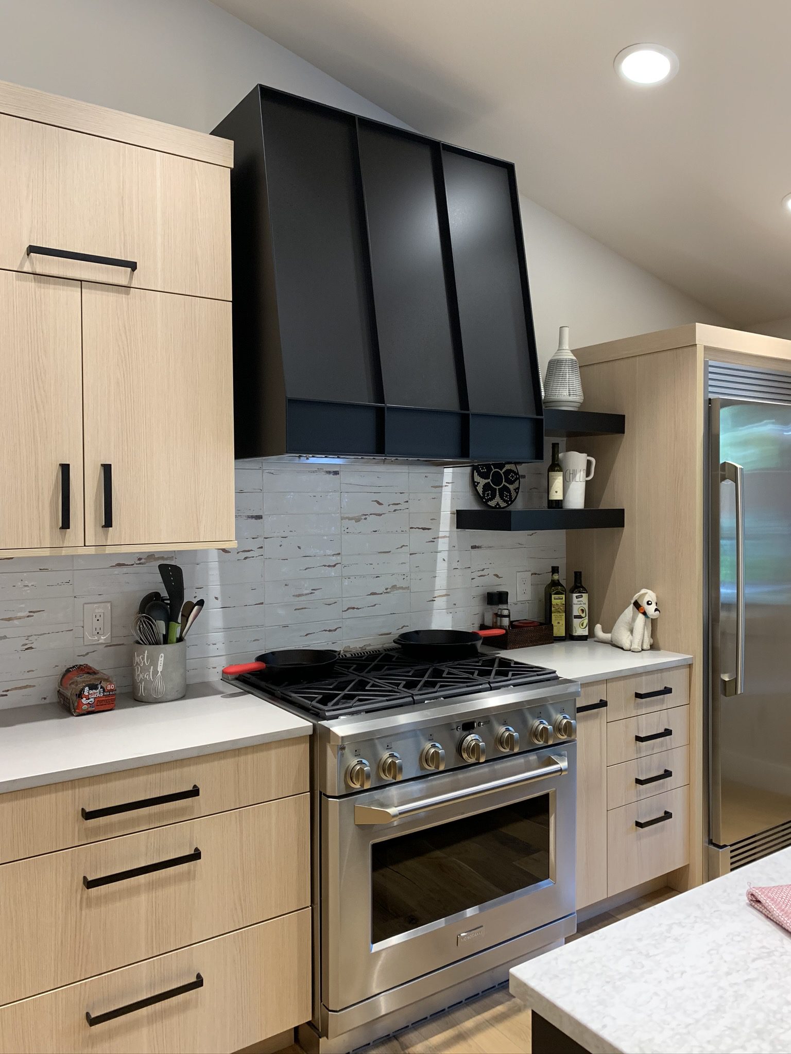 Modern kitchen with light wood cabinets, a stainless steel gas stove, black range hood, white backsplash, and open shelves holding decor items and spices.