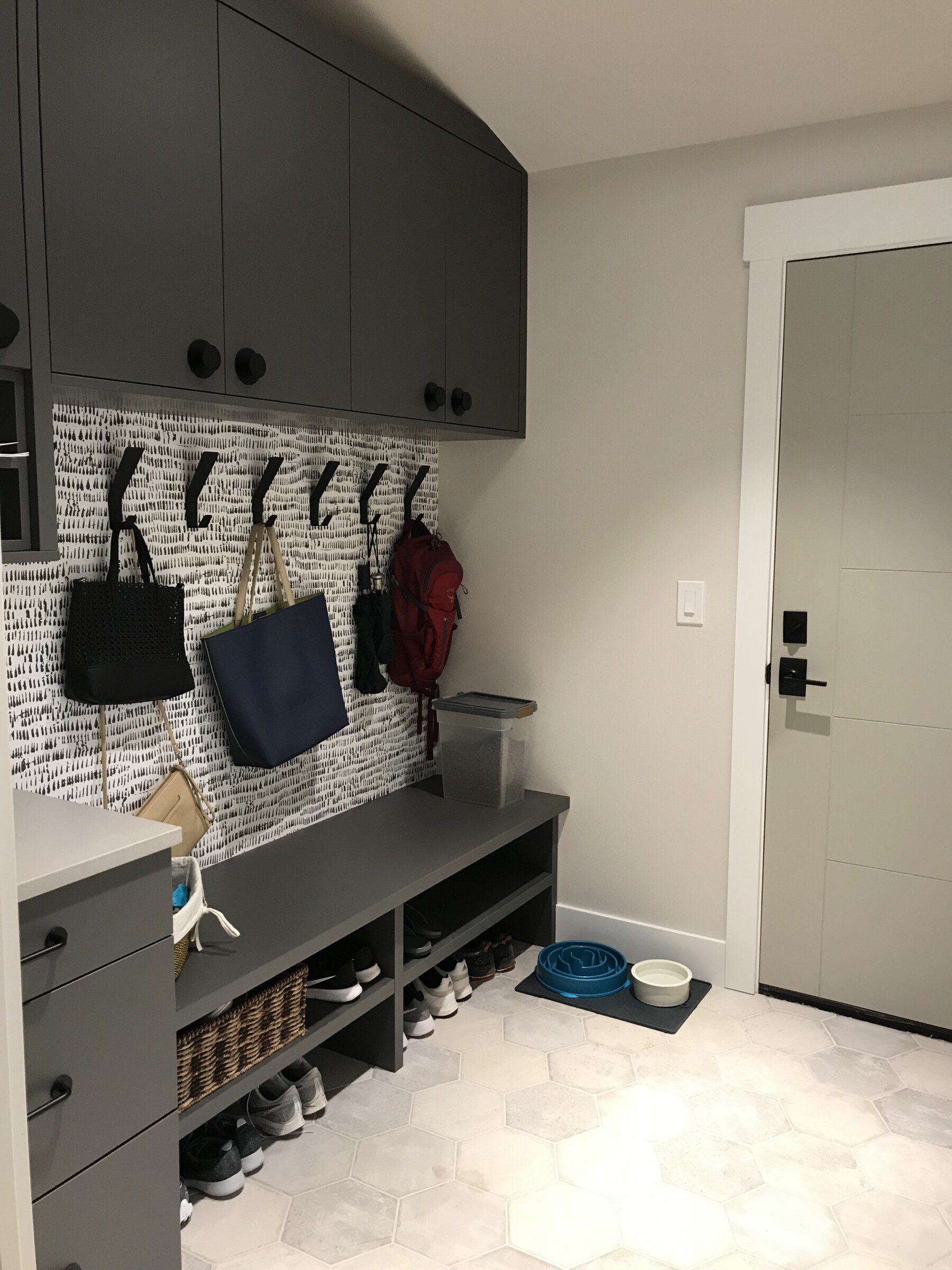 A mudroom with gray cabinets, black hooks holding bags, a bench with shoes underneath, a pet food bowl, and a light-colored door.